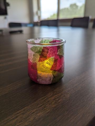 A small glass jar with a stained glass effect from green, maroon, and lavender tissue paper. It is glowing softly from an LED votive inside the jar. The jar sits on a dark wood table with a window in the background. 