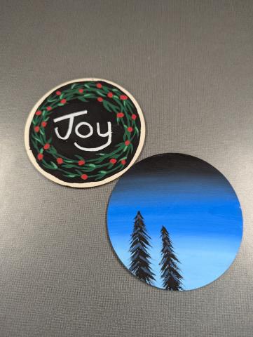 Two flat wooden circles that are painted. The circle on the top left has a black background, a green wreath design, and white letters spelling "Joy." The circle on the bottom right has a black to light blue ombre background with black tree silhouettes. 