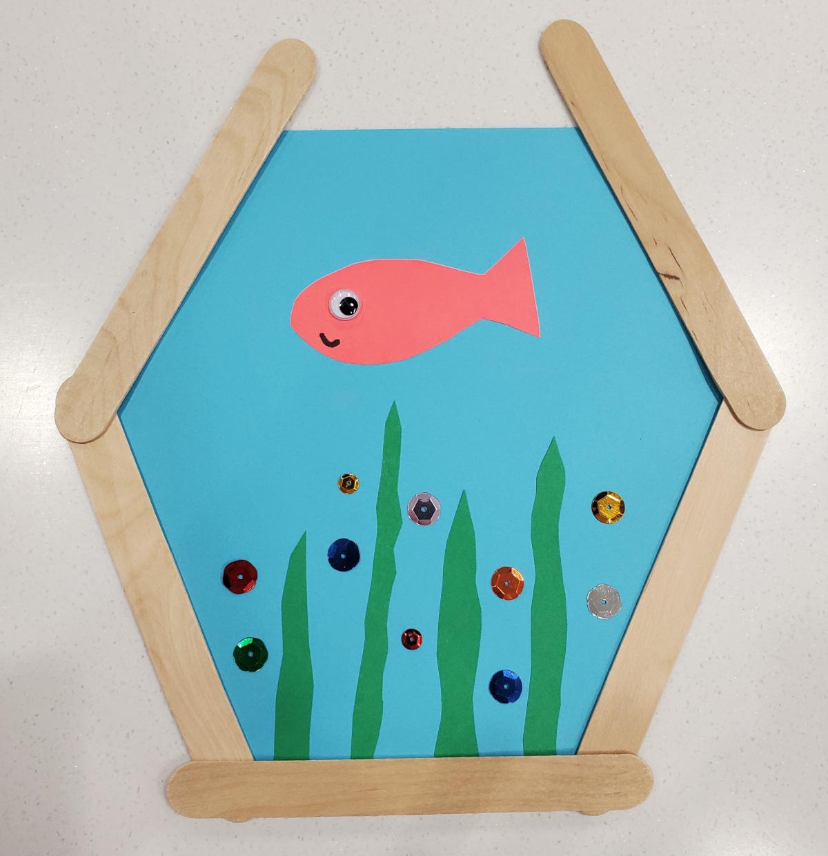 Photo of a fish bowl craft against a white background. The fish bowl craft is a blue paper with popsicle sticks around it in the shape of a fish bowl, glued to the blue paper is a cut out of a fish and some seaweed.